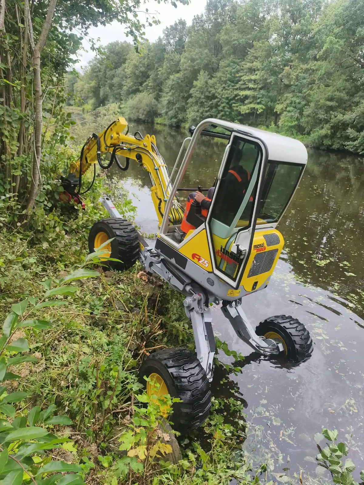 Menzi Muck A25 Schreitbagger im Einsatz zur Uferpflege Menzi Muck A25 Schreitbagger mit Mulchkopf bei der Landschaftspflege am Flussufer. Die Maschine steht mit ausgefahrenen Beinen auf unebenem Gelände und im Wasser.